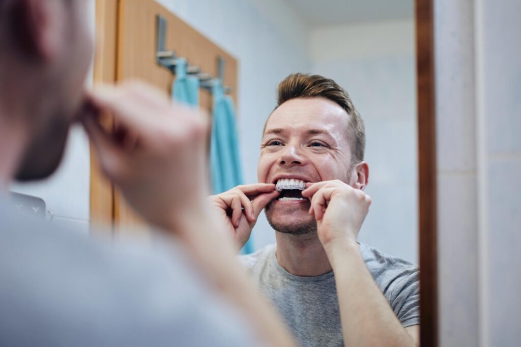 Man applying Invisalign aligners in bathroom mirror, demonstrating home care tips for orthodontic treatment.